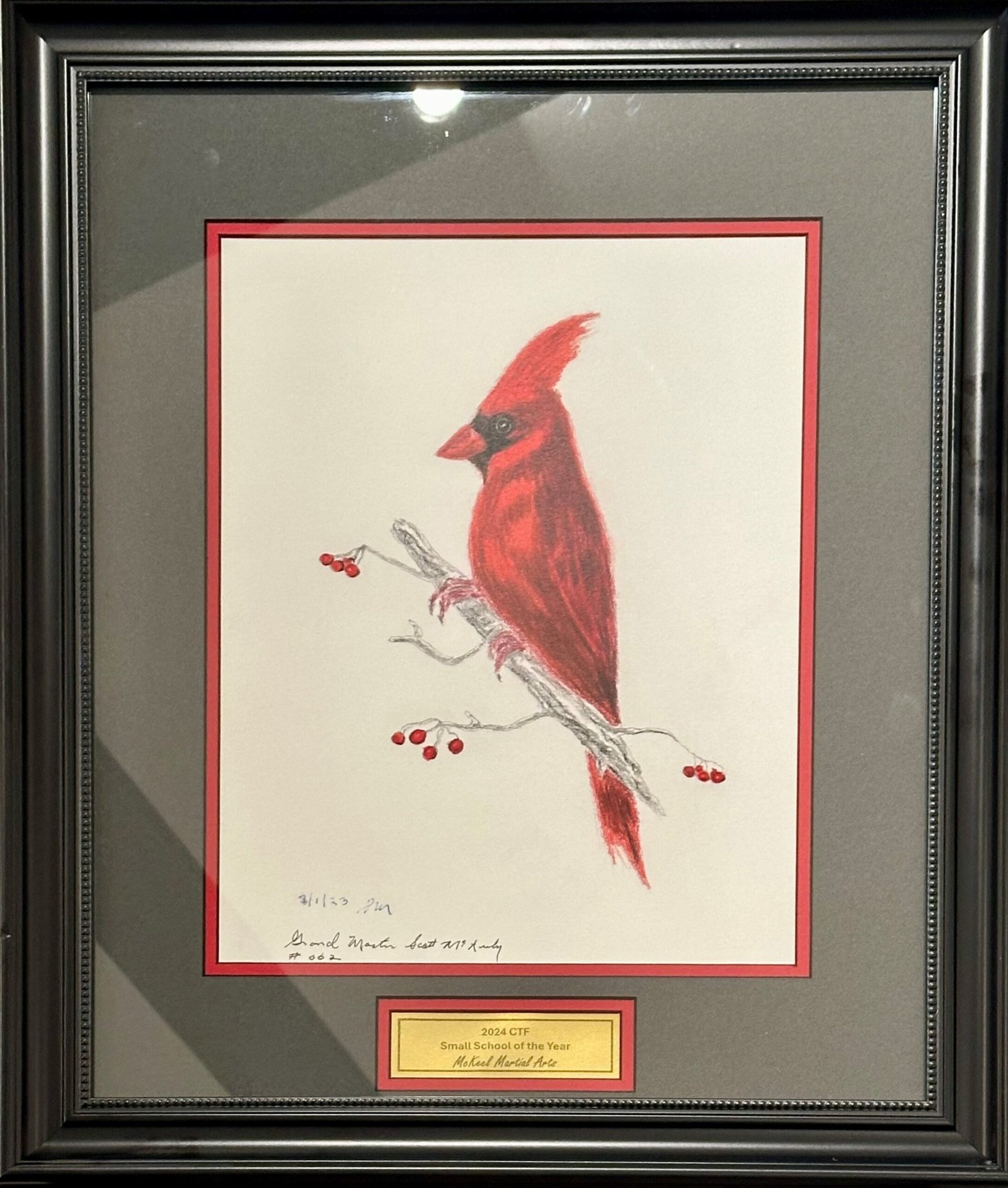 "A framed drawing of a red cardinal perched on a branch with red berries. The artwork is surrounded by a red and gray mat and encased in a black decorative frame.